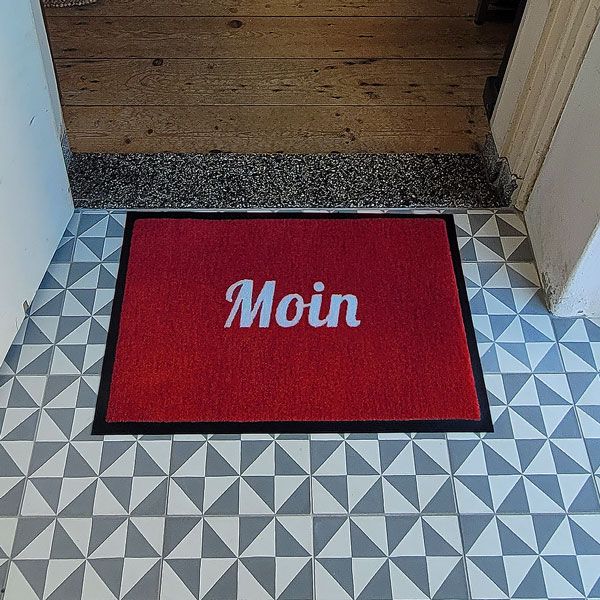 Fußmatte mit Aufschrift "Moin" in Rot mit weißer Schrift. Foto vor Küchentüre.