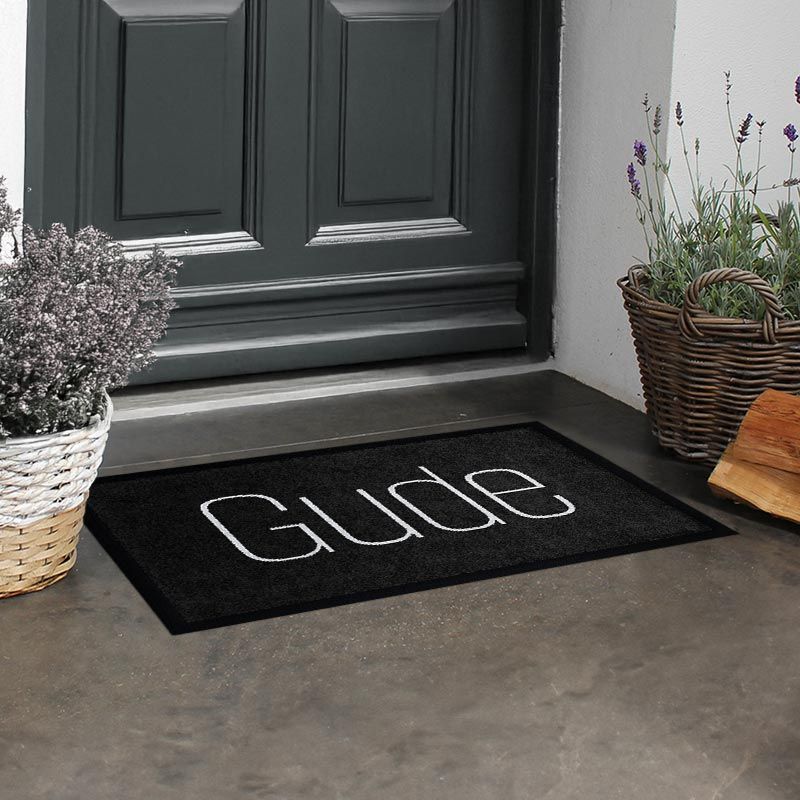 Waschbare Fußmatte von Glücksmatte mit dem Gruß "Gude" in moderner Schrift in Hellgrau auf Dunkelgrau. Beispielbild.